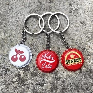 May include: Three keyrings with bottle top charms. The charms are red and white with different designs. One charm has a cherry design, another says "Nuka Cola", and the last one says "Sunset Sarsaparilla".