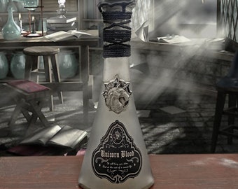 Peut inclure: Une bouteille en verre avec une étiquette noire qui indique "Sang de Licorne" et un emblème de tête de licorne argenté. La bouteille est attachée avec de la ficelle noire et a un sceau de cire noire en haut.