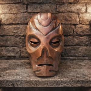 May include: A copper-colored mask with closed eyes and a stylized face. The mask has a smooth, metallic finish and is displayed against a textured stone surface and a brick wall background.