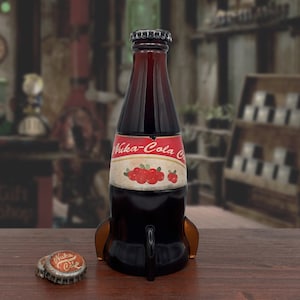 May include: A brown glass bottle shaped like a rocket with a red and white label that says "Nuka-Cola" and features red berries. The bottle is sitting on a wooden surface with a bottle cap on the surface.