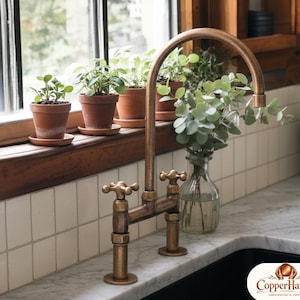 Könnte beinhalten: Ein kupferfarbener Wasserhahn im Vintage-Stil mit Kreuzgriffen. Der Wasserhahn ist auf einer Marmorarbeitsplatte neben einem schwarzen Waschbecken installiert. Eine Glasvase mit Grün steht in der Nähe des Wasserhahns. Kleine Topfpflanzen stehen auf einer Holzfensterbank.
