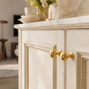 May include: A pair of polished gold-toned metal cabinet knobs with a rounded, ribbed design attached to off-white cabinet doors featuring decorative molding. The knobs have a reflective surface and a circular base plate mounted on the wood.