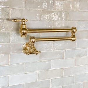 Op de afbeelding: Een aan de muur gemonteerde pannenvul-kraan met een gepolijste messing afwerking, voorzien van een dubbel scharnierende arm, twee ronde bedieningsknoppen en een uitloop. Het armatuur heeft een metallic goudkleurig oppervlak.