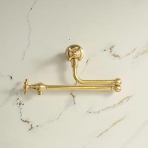 May include: A wall-mounted pot filler tap with a brushed gold finish. The fixture features a double-jointed articulating arm, a cross-handle control at the base, and a matching cross-handle at the spout end for water flow regulation.