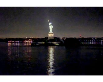 May include: The Statue of Liberty stands illuminated at night, positioned on a pedestal atop an island. The statue is bathed in bright white light, creating a distinct reflection on the dark water surface in the foreground.