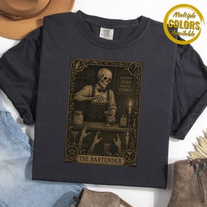 May include: Dark gray t-shirt featuring a detailed illustration of a skeleton bartender pouring a drink. The design includes the text "Happy Hour 6pm to Eternity" and "The Bartender". A gold circle with the text "Multiple Colors Available" is in the upper right.