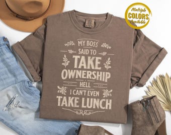 May include: A brown t-shirt with the text "MY BOSS SAID TO TAKE OWNERSHIP HELL I CAN'T EVEN TAKE LUNCH" in a white, decorative font. The shirt is displayed with blue jeans, a brown hat, and a pair of boots. The words "Multiple Colors Available" are in a yellow circle.