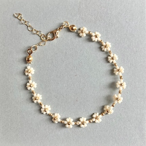 Peut inclure: Un bracelet délicat en chaîne dorée avec de petites perles blanches en forme de fleurs. Le bracelet est doté d'un fermoir à mousqueton.