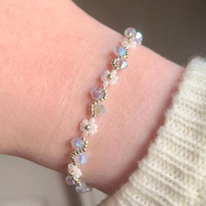May include: A delicate bracelet featuring iridescent crystal beads and small white flower-shaped beads. The bracelet is strung on a silver-colored wire and is approximately 7 inches in length. The bracelet is worn on a wrist.
