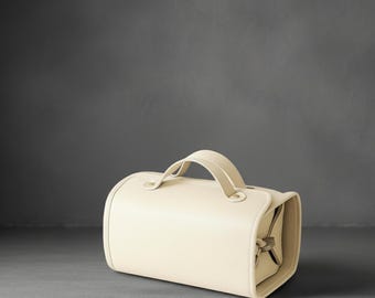 May include: Cream-colored, cylindrical travel bag with a curved handle. The bag has a rectangular base and a zippered compartment. The letters "AZ" are in the upper left corner.