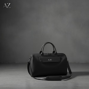 May include: Black duffel bag with a top handle and a shoulder strap. The bag has the initials "A.M" printed on the front. The bag is sitting on a gray surface.