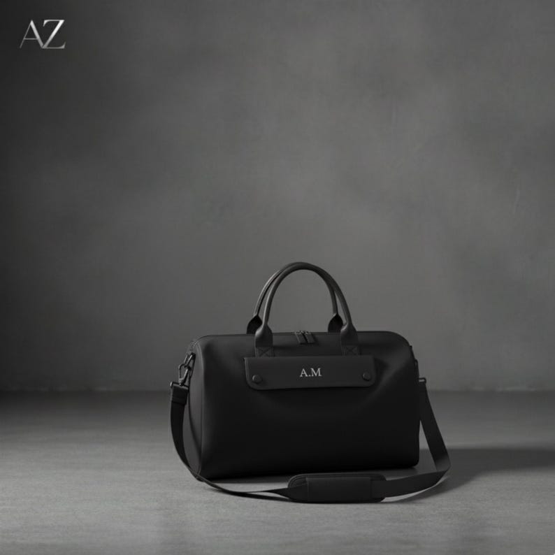 May include: Black duffel bag with a top handle and a shoulder strap. The bag has the initials "A.M" printed on the front. The bag is sitting on a gray surface.