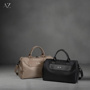 May include: Two personalized duffel bags. One is tan with the name "Lisa" and the other is black with "J.L.A." Both have top handles, side pockets, and shoulder straps. The bags are on a gray surface with a dark gray background.