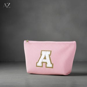 May include: A pink cosmetic bag with vertical stripes, featuring a large white letter "A" with a gold glitter outline. The bag has a green zip pull. The letters "AZ" are in the top left corner.