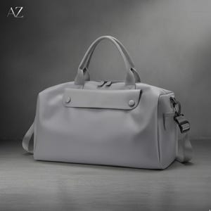 May include: A gray duffel bag with a top handle and a detachable shoulder strap. The bag has a front pocket with two button closures and a side pocket with a buckle. The letters "AZ" are in the top left corner.