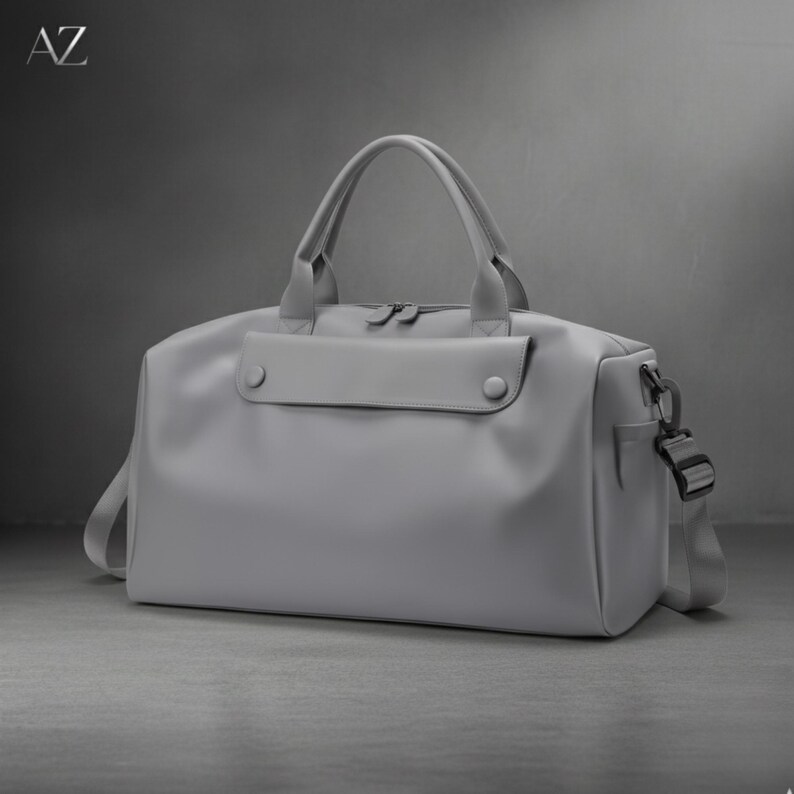 May include: A gray duffel bag with a top handle and a detachable shoulder strap. The bag has a front pocket with two button closures and a side pocket with a buckle. The letters "AZ" are in the top left corner.