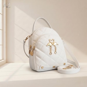 May include: White quilted backpack with gold-tone hardware. Features a top handle, adjustable shoulder strap, and a decorative gold swan emblem with dangling charms. The bag has a diamond pattern and gold accents.