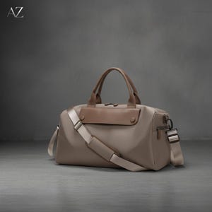 May include: A taupe duffel bag with brown leather accents, a top handle, and a detachable shoulder strap. The bag has a front flap and a zipper closure. The letters "AZ" are in the upper left corner.