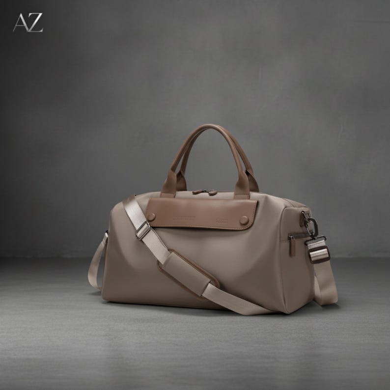 May include: A taupe duffel bag with brown leather accents, a top handle, and a detachable shoulder strap. The bag has a front flap and a zipper closure. The letters "AZ" are in the upper left corner.