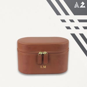 Puede incluir: Un neceser de viaje marrón, de forma ovalada, con cierre de cremallera y herrajes dorados. La funda tiene las iniciales "L.M" en letras doradas en la parte delantera. El fondo es blanco con un diseño geométrico en blanco y negro en la esquina superior derecha.