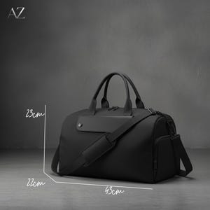 May include: A black duffel bag with a shoulder strap and handles. The bag is 43 cm long, 22 cm high, and 23 cm wide. It has a side pocket and a flap with a button. The letters "AZ" are in the top left corner.