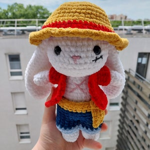 May include: White crocheted bunny wearing a yellow and red straw hat, a red vest, a blue denim skirt, and a yellow belt.