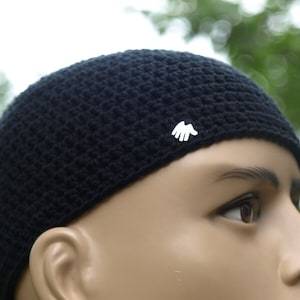 Könnte beinhalten: Schwarze Häkelmütze mit einem kleinen weißen Handsymbol an der Seite.