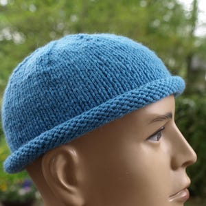 Könnte beinhalten: Eine blaue Strickmütze mit abgerundeter Krone und einem dicken, gerollten Rand. Die Mütze besteht aus strukturiertem Garn mit einem sichtbaren glatten Rechtsstrickmuster am Hauptteil und einem kraus rechts gestrickten Rand.