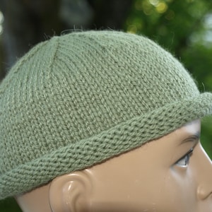May include: A sage green knitted beanie hat featuring a rounded crown and a thick, rolled brim. The hat is constructed with a vertical ribbed stitch pattern on the main body and a garter stitch texture on the folded edge, providing a simple, clean look.