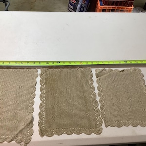 May include: Three beige crocheted placemats with a scalloped edge. The placemats are laid out on a white table with a measuring tape.
