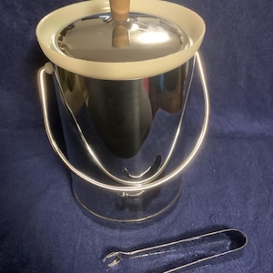 May include: A stainless steel ice bucket with a lid and a wooden handle. The bucket has a silver handle and is accompanied by a pair of silver ice tongs. The ice bucket and tongs are on a blue surface.