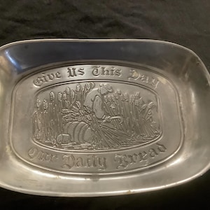 May include: A rectangular pewter tray with rounded corners. The tray features an embossed scene of a person harvesting wheat, with the words "Give Us This Day Our Daily Bread" above and below the image. The tray has a slightly tarnished appearance.