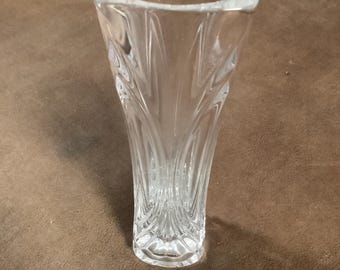 May include: A clear glass vase with a square top and a flared, textured body. The vase has a decorative pattern that runs vertically along its sides. The vase is empty and sits on a brown surface.