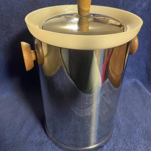 May include: A stainless steel ice bucket with a lid and wooden handle. The bucket has a white rim and two wooden handles on the sides. The lid also has a wooden handle. The bucket is cylindrical and reflective.