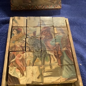 May include: Vintage wooden block puzzle featuring illustrations of people and animals in a classic style. The blocks are arranged in a wooden tray, with a matching lid visible in the background. The images on the blocks are in various colors.