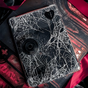May include: A black phone case with a white spiderweb design, a black heart, and a black pop socket. The case is on a dark background with red ribbon.