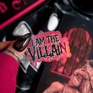 Op de afbeelding: Een sticker met de tekst "I AM THE VILLAIN" in zwart gotisch lettertype, omlijnd in wit en rood. De sticker wordt voor een donkere achtergrond met andere items gehouden. Het logo The Pumpkin Pot is zichtbaar.