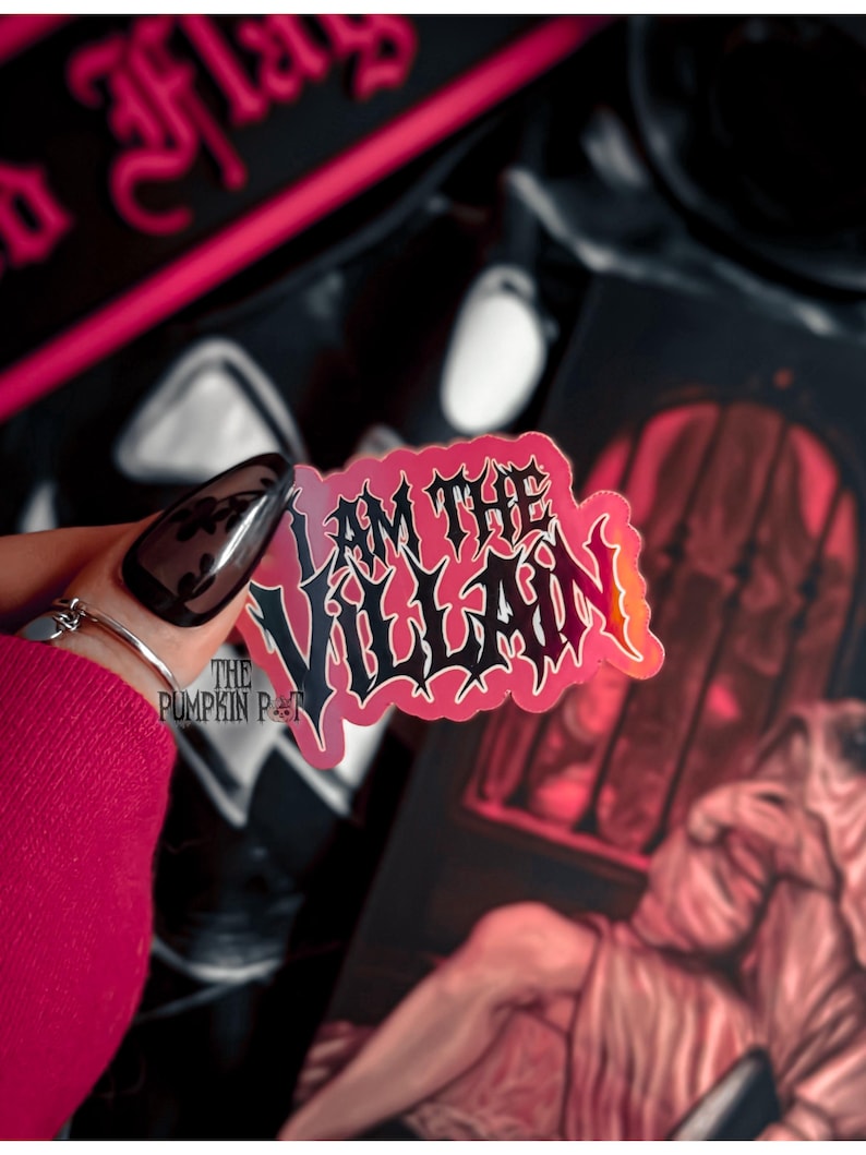 Op de afbeelding: Een sticker met de tekst "I AM THE VILLAIN" in zwart gotisch lettertype, omlijnd in wit en rood. De sticker wordt voor een donkere achtergrond met andere items gehouden. Het logo The Pumpkin Pot is zichtbaar.
