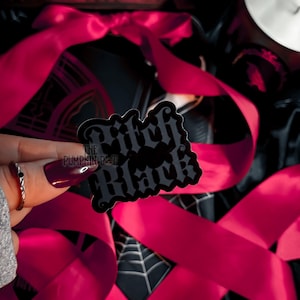Puede incluir: Una pegatina negra y gris con las palabras "Witch Black" en una fuente gótica. La pegatina se sostiene en una mano con esmalte de uñas rojo oscuro y un anillo plateado. Se ven cintas rosas brillantes y un fondo negro con un diseño de telaraña.