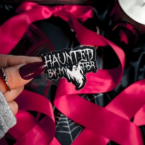 Puede incluir: Un pin negro y blanco con el texto "HAUNTED BY MY TBR" y una ilustración de un fantasma. El pin se sostiene frente a un fondo de cinta rosa brillante y un fondo negro con un diseño de telaraña.