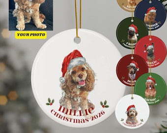 May include: A round white ceramic Christmas ornament featuring a custom illustration of a brown spaniel wearing a red Santa hat. The text MILLIE CHRISTMAS 2026 is printed below the dog. A gold hanging string is attached to the top of the ornament.