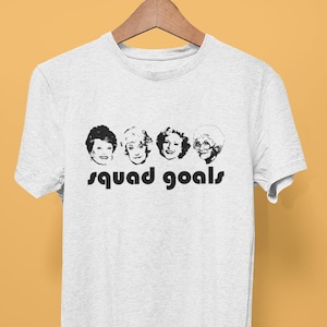 May include: A light grey t-shirt with a black and white graphic of four women's faces and the words "squad goals" in black. The t-shirt is displayed on a wooden hanger against a yellow background.