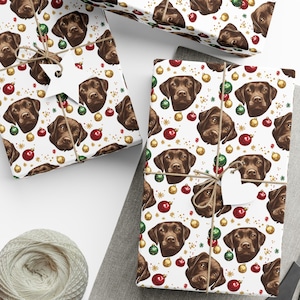 May include: White wrapping paper featuring a repeating pattern of brown Labrador retriever faces, red and green Christmas ornaments, and gold star accents. The paper is tied with brown twine and includes white star and heart-shaped gift tags.