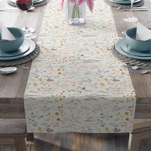 May include: A cream-coloured floral table runner featuring a delicate wildflower pattern in yellow, purple, and pink. The runner is laid on a wooden table, set with place settings, wine glasses, and a vase of pink lilies.
