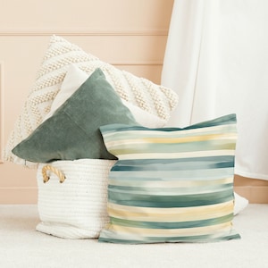 May include: Three decorative throw pillows in a white wicker basket. The pillows are a white textured pillow, a dark green velvet pillow, and a blue and green striped pillow.