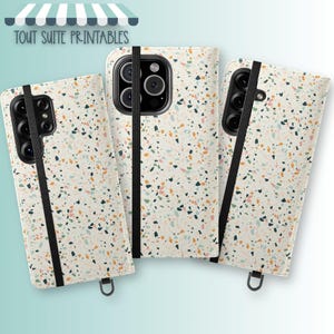 May include: Three phone cases with a cream-colored terrazzo pattern, featuring black elastic bands and a black camera module. The cases have a variety of colorful flecks. The text "TOUT SUITE PRINTABLES" is at the top.