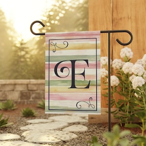 May include: A garden flag with a watercolor stripe design in shades of pink, yellow, and green. The letter "E" is in the center of the flag in black.