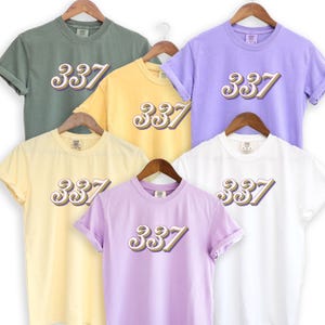 Puede incluir: Cinco camisetas con el número 337 impreso en una fuente retro y colorida. Las camisetas son de varios colores, incluyendo verde, amarillo, morado y blanco.