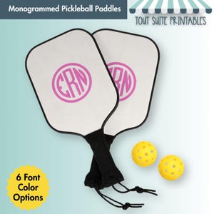 Könnte beinhalten: Zwei weiße Pickleball-Schläger mit schwarzen Griffen und einem rosa Monogramm. Zwei gelbe Picklebälle sind ebenfalls abgebildet. Der Text "Monogrammed Pickleball Paddles" steht oben.