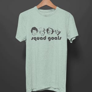 May include: A pale green t-shirt featuring a black graphic of four women's faces and the words "squad goals" printed underneath. The t-shirt is suspended on a wooden hanger against a dark grey backdrop.